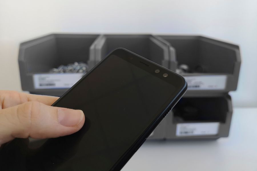Im Vordergrund eine Hand, die ein schwarzes Smartphone mit ausgeschaltetem Bildschirm hält. Im Hintergrund sind drei graue Aufbewahrungsbehälter mit kleinen Gegenständen darin auf einer weißen Oberfläche platziert.